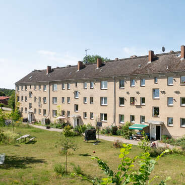 A three-story beige apartment building with a sloped roof and several chimneys. The building overlooks a grassy courtyard with benches, plants, and outdoor equipment.