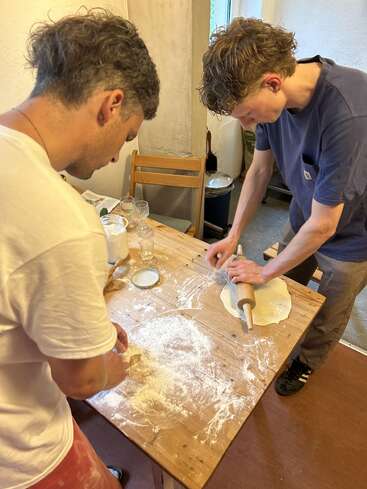 Two people in a cozy kitchen are making dough. One uses a rolling pin, while the other adds flour. The wooden table is scattered with flour.