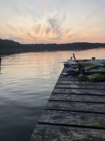 A serene lakeside at sunset, wooden dock extends over calm water, person swimming, clothes and bags scattered, dramatic clouds painted across the softly glowing evening sky.