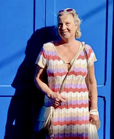 Eine lächelnde Frau steht vor einer leuchtend blauen Wand. Sie trägt ein buntes Kleid mit Zickzackmuster, hat eine Sonnenbrille auf dem Kopf und hält eine Umhängetasche sowie eine weiße Tragetasche in der Hand.