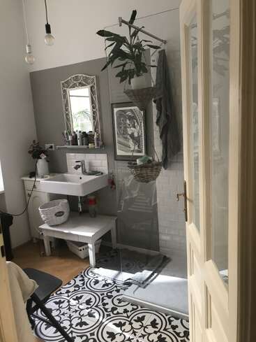 Dieses gemütliche Badezimmer besticht durch gemusterte Bodenfliesen, ein weißes Waschbecken, einen dekorativen Spiegel, hängende Pflanzen, Kunstwerke und moderne Beleuchtung. Ein Duschbereich und Regale sorgen für funktionalen Stil.