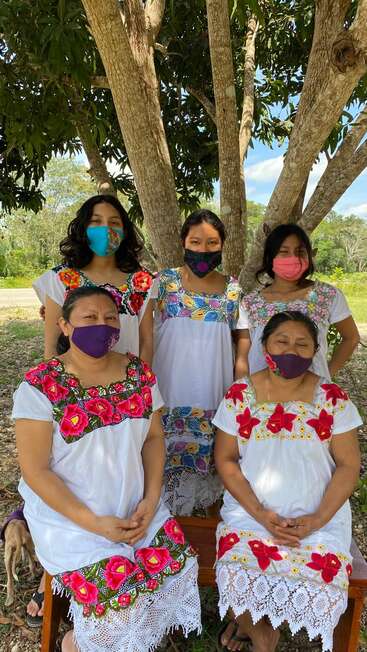 Cinco mujeres con coloridos vestidos tradicionales bordados y máscaras faciales se sientan y permanecen de pie bajo un gran árbol al aire libre, posando juntas en un día soleado.