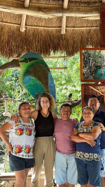 Cinco personas sonrientes permanecen juntas bajo un tejado de paja, con un colorido mural de pájaros y un bosque de fondo, irradiando felicidad y sensación de unión.
