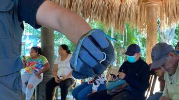 Una persona con un guante azul sostiene una pequeña serpiente. Varias personas sentadas al fondo, algunas utilizando teléfonos, bajo un tejado de paja en un entorno al aire libre.
