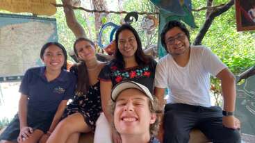 Cinco amigos están sentados juntos, sonriendo alegremente para una foto de grupo. Les rodea una luz brillante y natural, con mapas y vegetación visibles al fondo, creando un ambiente alegre.