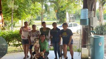 Seis personas y un perro posan juntos en un patio soleado con árboles verdes, plantas y un coche blanco de fondo junto a una fuente de agua.