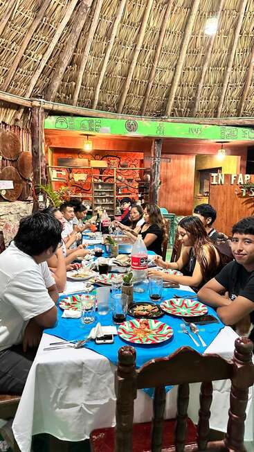 Un grupo de personas se reúne alrededor de una mesa decorada en un restaurante rústico con tejado de paja, disfrutando de la comida, la bebida y la conversación en un ambiente cálido y acogedor.
