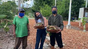 Tres personas están de pie al aire libre, todas con máscaras faciales. Dos mujeres sostienen cestas llenas de grandes cangrejos, mientras un hombre permanece de pie junto a ellas, rodeado de vegetación y árboles.