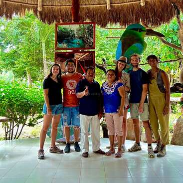 Un groupe de sept personnes souriantes, posant ensemble sous un toit de chaume dans un cadre tropical avec une végétation luxuriante, des œuvres d'art et deux photos en arrière-plan.