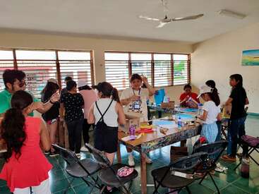 Un aula llena de niños y algunos adultos. Se dedican a actividades artísticas y manuales, utilizando papel, cajas y pegamento, creando un ambiente animado y creativo.