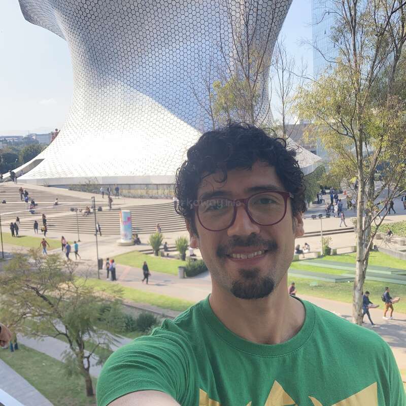 A smiling man with glasses takes a selfie outdoors. In the background, there’s a futuristic metallic building, trees, steps, and people walking in a sunny setting.