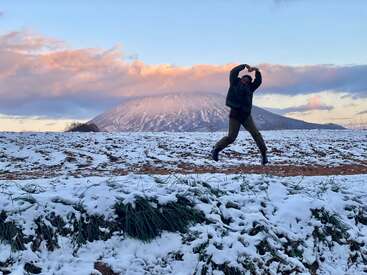 Uma pessoa pula em um campo nevado, fazendo um formato de coração com as mãos, com uma montanha coberta de neve ao fundo, sob um céu azul com nuvens cor-de-rosa.