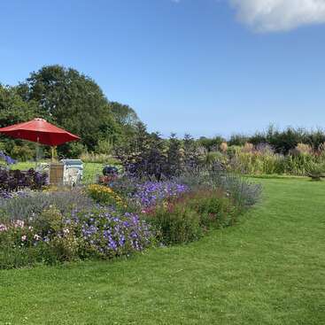 The image depicts a serene garden scene with a vibrant flowerbed, lush green grass, and a red umbrella, set against a clear blue sky.