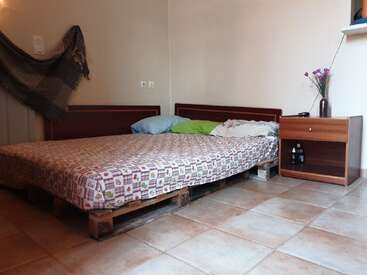 Ein einfaches Schlafzimmer mit einem Palettenbett, bunten Kissen, einer gemusterten Tagesdecke, einem hölzernen Nachttisch mit Flaschen, einem Fliesenboden, einem Wandteppich und einer Vase mit lila Blumen.