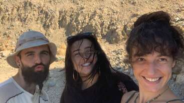 Three friends pose outdoors on a sunny day, smiling and enjoying themselves in a rocky, desert-like landscape. Wind blows their hair playfully across their faces.