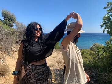 Two people, one wearing sunglasses, dance joyfully near the seaside. Dry bushes and green plants surround them, with beautiful blue ocean and clear sky in the background.