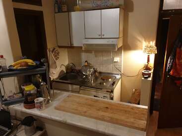 Una pequeña cocina acogedora con una tabla de cortar de madera, platos secándose, cocina eléctrica, armarios superiores, lámpara de calor, frutero y varios utensilios de cocina sobre la encimera.