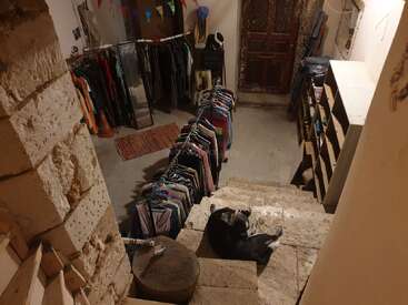 Una acogedora tienda de segunda mano en un sótano con escalones de piedra, un perro negro tumbado, ropa de colores en percheros, estanterías, colorines festivos y una rústica puerta de madera.