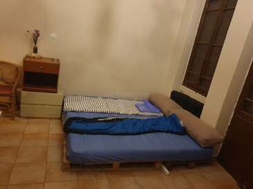 Esta habitación cuenta con una sencilla cama hecha de palés con ropa de cama azul, una manta a rayas, una ventana de madera, un pequeño armario, una silla y un jarrón.
