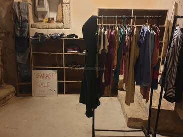 Un acogedor montaje de venta de garaje interior con percheros de ropa de colores, estanterías con vaqueros y accesorios, y un cartel hecho a mano en el que se ofrecen artículos de aportación gratuita.