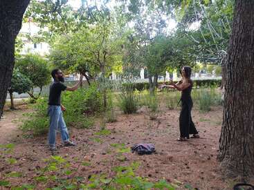 Dos personas están de pie en una zona boscosa y sombreada haciéndose gestos con las manos. Árboles y arbustos los rodean, con una chaqueta tirada en el suelo cerca.