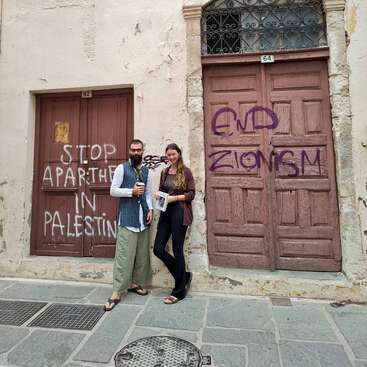 Deux personnes se tiennent devant de vieilles portes en bois portant des graffitis politiques indiquant "STOP APARTHEID IN PALESTINE" et "END ZIONISM" dans une rue abîmée par les intempéries.