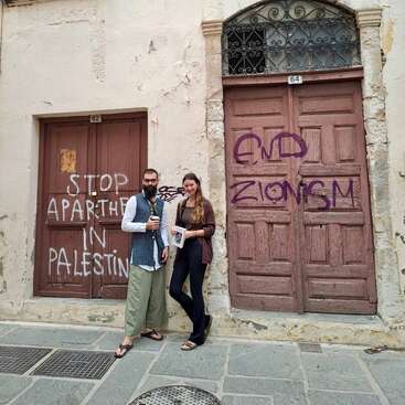 Two people stand in front of old wooden doors with political graffiti reading "STOP APARTHEID IN PALESTINE" and "END ZIONISM" on a weathered street.