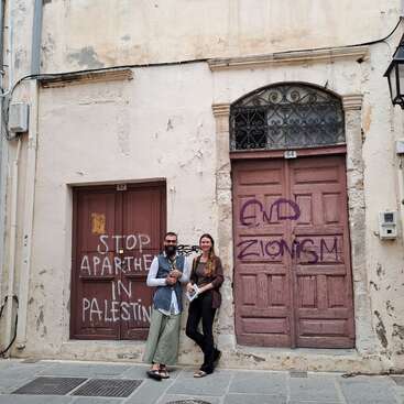 Deux personnes se tiennent devant un vieux bâtiment dont les portes sont recouvertes de graffitis disant « STOP À L'APARTHEID EN PALESTINE » et « FIN AU SIONISME ». Il y a un pot de fleurs et un lampadaire à proximité.