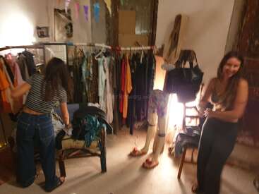 Dos mujeres se encuentran en una habitación acogedora y cálidamente iluminada, llena de percheros, prendas colgadas, un maniquí, bolsos y banderines festivos, lo que crea un ambiente animado y alegre.