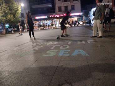 Abends versammeln sich die Leute auf einem Stadtplatz mit beleuchteten Geschäften. Jemand schreibt mit bunter Kreide die Worte „VOM FLUSS ZUM MEER“ auf den Bürgersteig.
