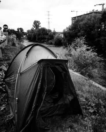 Ein Schwarz-Weiß-Foto zeigt ein Zelt, das in der Nähe eines Flussufers aufgeschlagen wurde, mit Bäumen, Baukränen und Gebäuden im Hintergrund – es vermittelt den Eindruck von Camping in der Stadt.