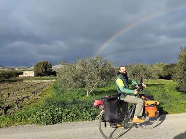 Ein Mann auf einem voll beladenen Tourenrad hält auf einer Landstraße an. Grüne Felder, Bäume und ein Regenbogen unter bewölktem Himmel sorgen für einen friedlichen, malerischen Moment.