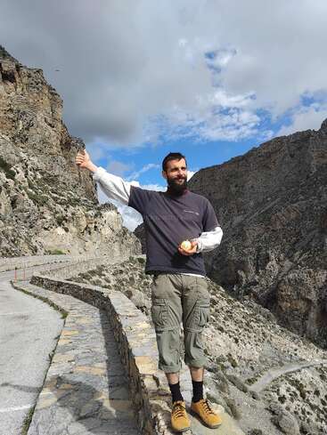 A man stands on a winding mountain road, holding an orange, smiling, and giving a thumbs up. Rugged rocky cliffs and cloudy blue sky surround him.