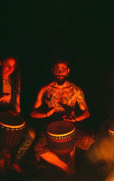 Two people are illuminated by warm firelight, playing hand drums. The central figure is shirtless with body paint and tattoos, creating a mystical, tribal ambiance at night.