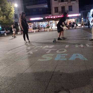 People gather at night in a city square with illuminated shops. Someone writes colorful chalk words on the pavement that read “FROM THE RIVER TO THE SEA.”