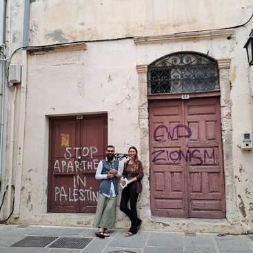 Two people stand in front of old buildings with graffiti reading “STOP APARTHEID IN PALESTINE” and “END ZIONISM.” The atmosphere is peaceful, featuring political messages.