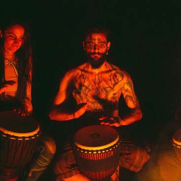 Two people are illuminated by warm firelight, playing hand drums. The central figure is shirtless with body paint and tattoos, creating a mystical, tribal ambiance at night.