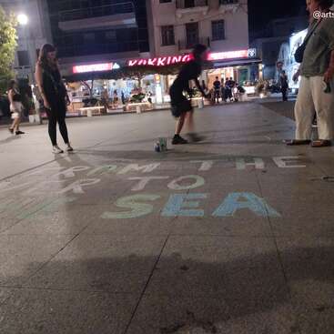 Le soir, les gens se rassemblent sur une place de la ville bordée de boutiques illuminées. Quelqu'un écrit à la craie colorée sur le trottoir : « DE LA RIVIÈRE À LA MER ».