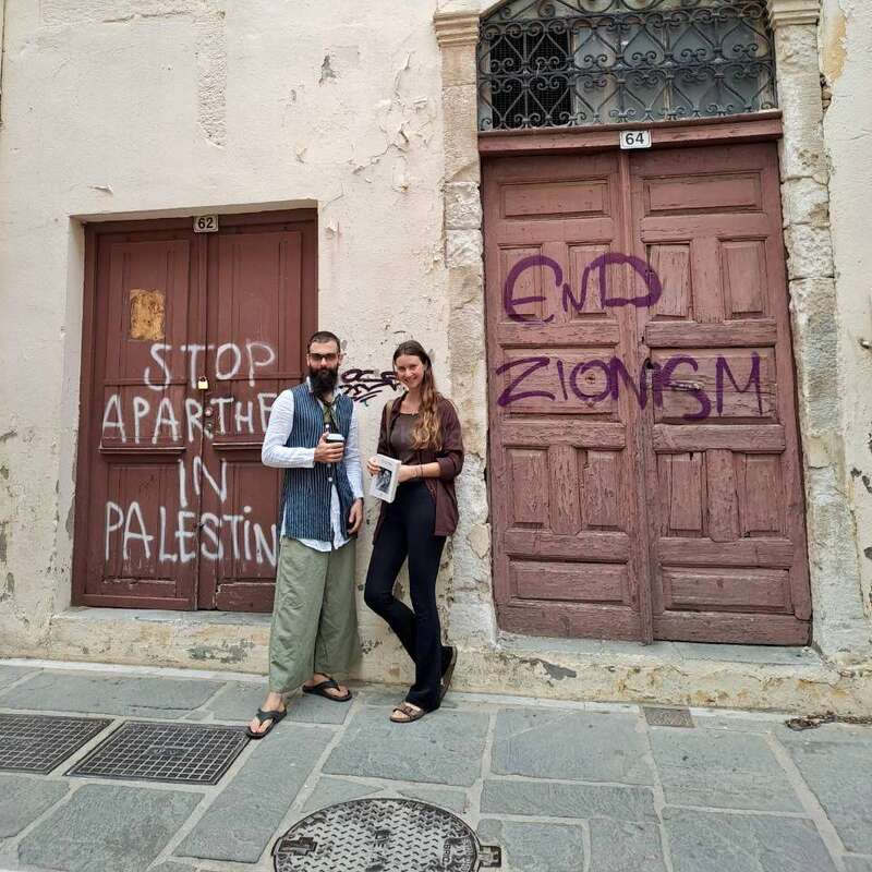 Dos personas ante unas viejas puertas de madera con pintadas políticas en las que se lee "STOP AL APARTHEID EN PALESTINA" y "FIN AL SIONISMO" en una calle desgastada.