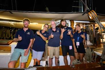 Cinq personnes se tiennent debout sur un voilier, de nuit, le sourire aux lèvres, faisant le signe du pouce levé et le « shaka ». Elles portent des chemises bleu marine assorties et profitent ensemble d'une aventure en mer.