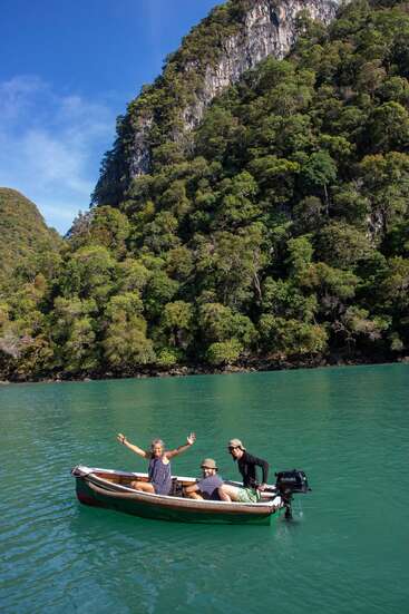 Trois personnes se trouvent à bord d'un petit bateau à moteur sur des eaux turquoise, entourées de falaises recouvertes d'une jungle luxuriante, profitant d'une journée ensoleillée, le sourire aux lèvres et les bras grands ouverts.