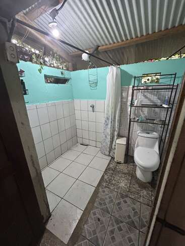 Dieses Bild zeigt ein einfaches Badezimmer mit türkisfarbenen Wänden, weißen Fliesen, einem Duschbereich, einer Toilette, einem Duschvorhang, offenen Regalen und einem Wellblechdach.