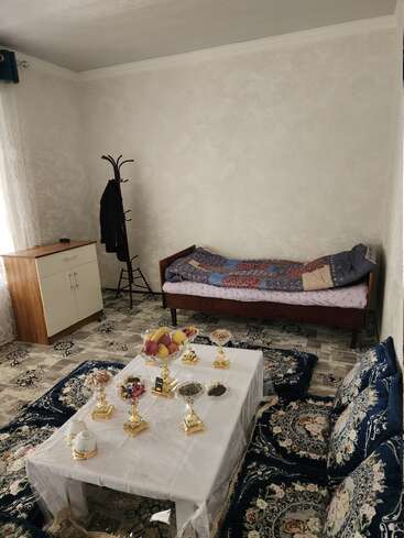 Une chambre confortable avec un lit simple, un porte-manteau et une armoire. Une table basse est dressée avec des fruits et des collations, entourée de coussins aux motifs décoratifs.