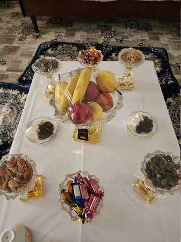 Une table magnifiquement dressée avec des fruits, des noix, des chocolats et des bonbons dans des coupes en verre décoratives, posées sur une nappe blanche. Des tapis aux motifs traditionnels entourent la table.