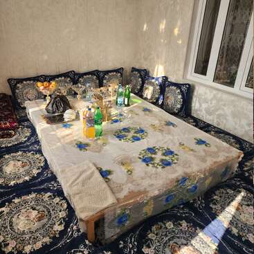 Une salle à manger traditionnelle avec des coussins et un tapis à motifs floraux bleus, une table basse recouverte d'une nappe décorative, des collations, des boissons, des fruits et la lumière étincelante du soleil.