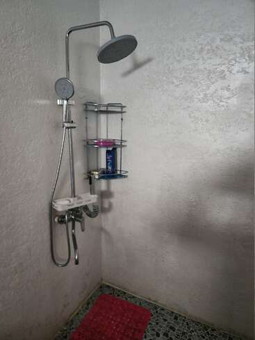 Cette image montre un coin salle de bain simple avec une douche murale, une étagère métallique pour les articles de toilette, des murs texturés, un sol à motifs et un tapis de bain rouge.