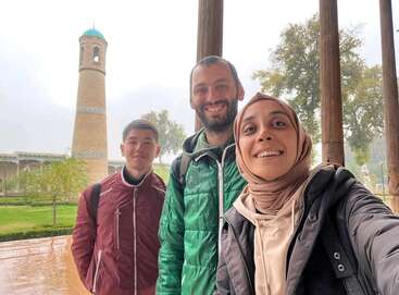 Trois amis posent joyeusement pour un selfie à côté de colonnes en bois, avec un minaret historique, des jardins verdoyants et des arbres visibles en arrière-plan par une journée nuageuse.