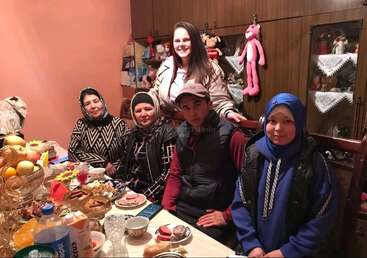 Un groupe de cinq personnes est assis autour d'une table avec de la nourriture, des boissons et des desserts. Une femme se tient derrière eux, tout le monde sourit dans une pièce confortable et chaleureusement éclairée.