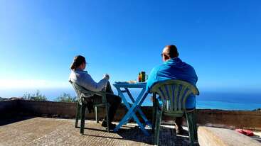 Deux personnes sont assises à une petite table en terrasse et partagent un repas. Face à l'océan d'un bleu magnifique sous un ciel dégagé, elles se détendent paisiblement tout en discutant.
