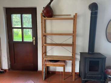 Ein gemütlicher Raum hat eine Holztür, ein leeres Bücherregal aus Holz, ein Musikinstrument drauf, einen Holzofen, eine Bank und Fliesenboden.